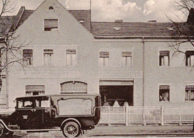Historische Aufnahme des damaligen Firmengebäudes in Guben mit Bestattungsfahrzeug vor dem Haus.